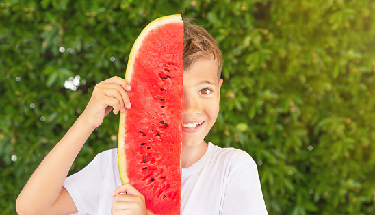 Happy child (boy) with a red juicy, tasty watermelon. Caucasian kid smiling (laughing) and having fun. Concept of healthy food, happy childhood, summer vacation. Nature green background. Copy space.