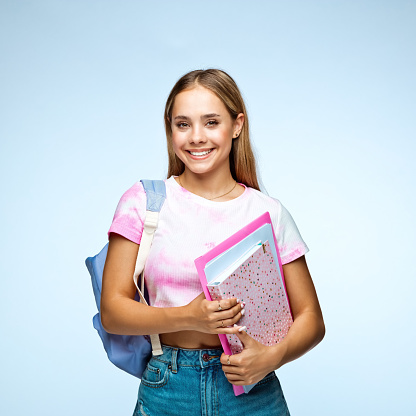 Portrait of smiling teenage girl with backpack and files standing against blue background.