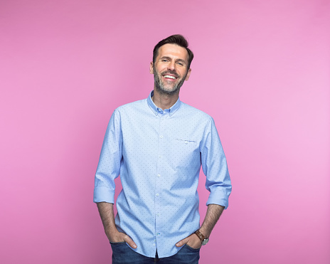 Portrait of confident mid adult man standing with hands in pockets against pink background