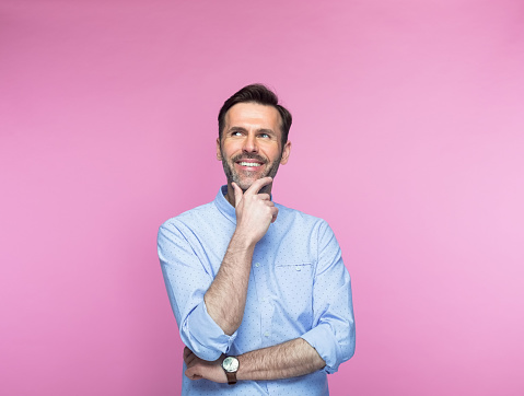 Thoughtful mid adult man with hand on chin looking up against pink background