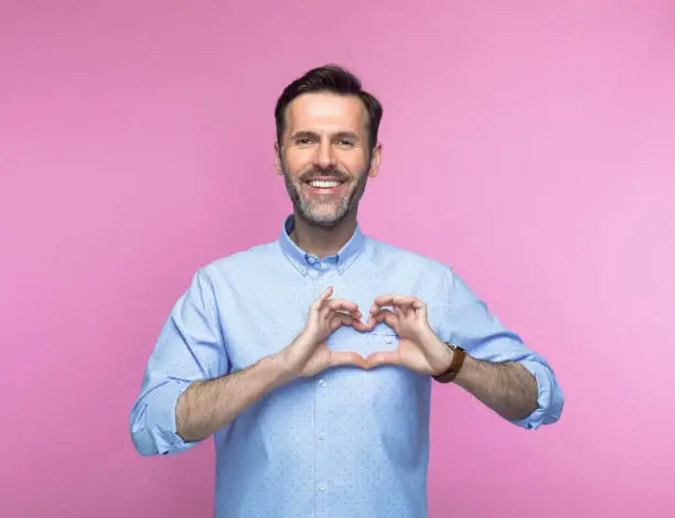 Portrait of smiling mid adult man in shirt gesturing heart sign against pink background