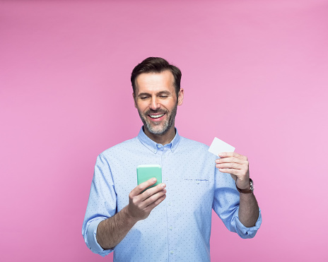 Happy man with smart phone holding credit card against pink background