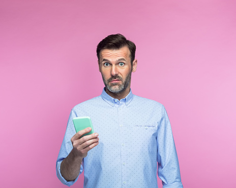 Portrait of shocked mid adult man with smart phone standing against pink background