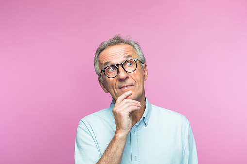 Confused retired elderly man with hand on chin looking away while thinking against pink background