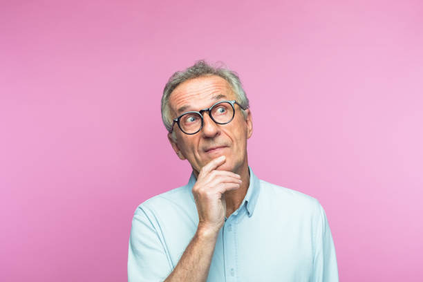 Confused retired man looking away while thinking Confused retired elderly man with hand on chin looking away while thinking against pink background confusion raised eyebrows human face men stock pictures, royalty-free photos & images