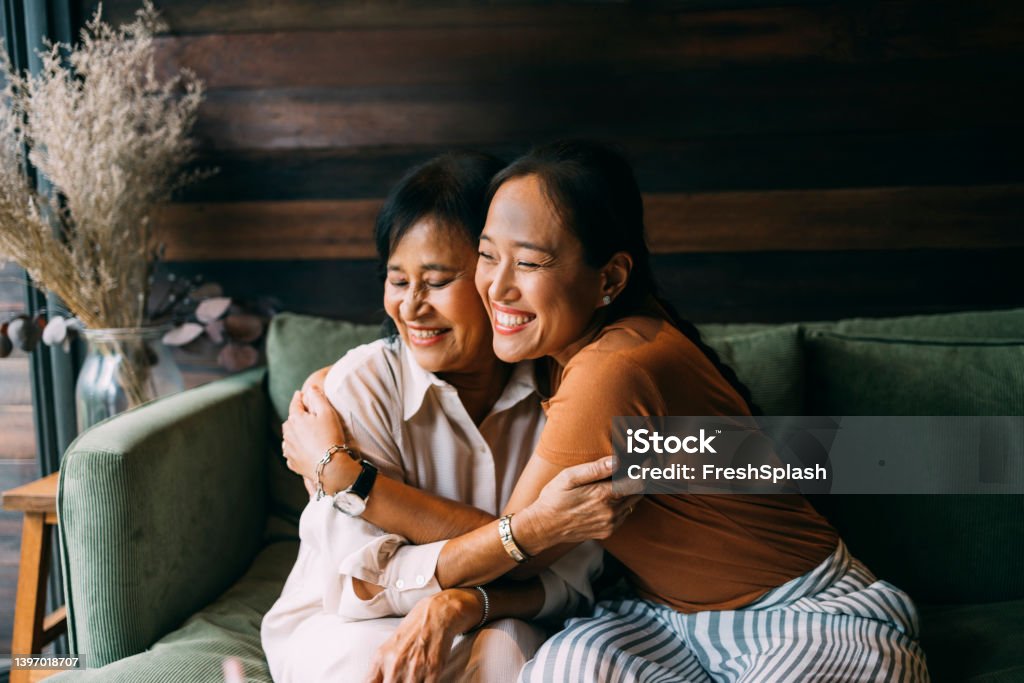 Mother And Daughter Hugging Each Other Asian mother and her daughter sitting on the sofa in their living room, spending time together. Mother Stock Photo