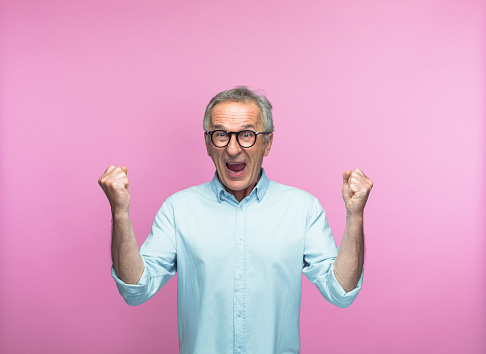 Portrait of excited senior man shouting while gesturing with fists against pink background