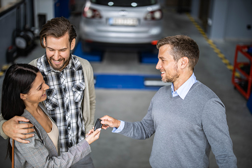Happy salesman giving car keys to young couple in auto repair shop.