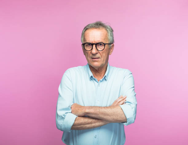 Serious retired senior man with arms crossed Portrait of serious retired senior man standing with arms crossed over pink background rolled up sleeves stock pictures, royalty-free photos & images