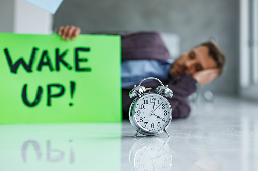 Close up of an alarm clock on the floor with a businessman in the background waking up. Copy space.