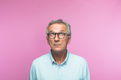 Thoughtful retired elderly man wearing eyeglasses looking up with raised eyebrows against pink background