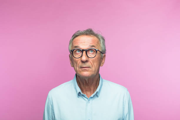 Thoughtful retired elderly man looking up Thoughtful retired elderly man wearing eyeglasses looking up with raised eyebrows against pink background Confusion stock pictures, royalty-free photos & images