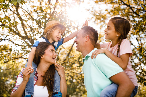 Young happy family having fun in spring day at the park. Mother is carrying son on shoulders while father is piggybacking daughter. Kids are giving each other high-five.