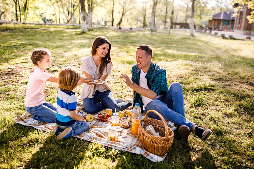 A happy vegan family on a picnic, mother plays the guitar and father plays with the children, fruit and vegan food on the picnic blanket.