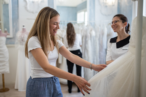 Caucasian female customer, a future bride, at the bridal shop with a help of saleswoman choosing her wedding dress