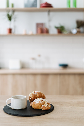 Fresh homemade croissant and coffee cup on black plate. Delicious food and hot drink on wooden table. Morning breakfast at clean countertop against blurred kitchen cabinet on background