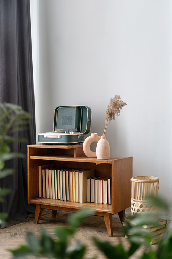 Concept of vintage objects and old fashionable decor in cozy living room. Vertical view of retro style vinyl player stand in apartment against white copy space wall on wooden commode with paper books