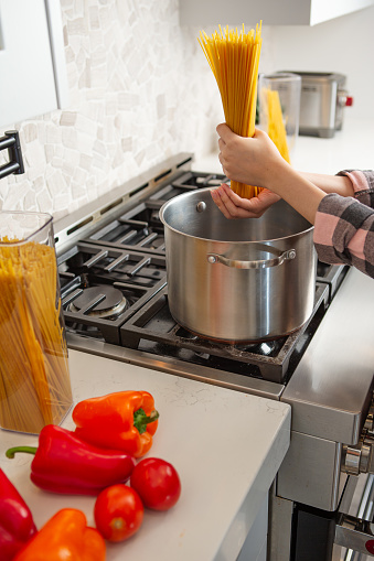 Female hand holding a handful of raw pasta noodle in pot on gas stove in kitchen.