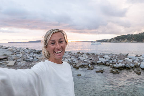 joven junto al mar toma una foto selfie para recuerdos. sonríe a la cámara feliz de estar de vacaciones - blond hair overcast sun sky fotografías e imágenes de stock