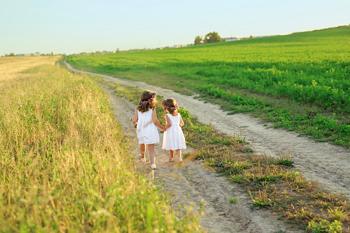 Children run hand in hand in the fresh air among fields and meadows on a narrow sandy path. Girls in dresses and with wreaths of flowers in their hair at sunset in nature