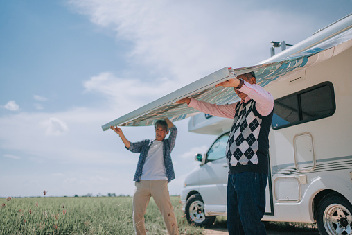 2 asian chinese senior man setting up awning canopy of campervan motor home during day time beside rice paddy field