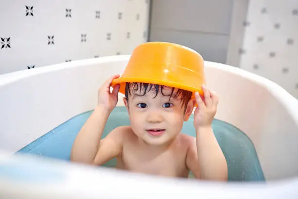 One asian boy wearing washbowl on head and playing at bathtub.