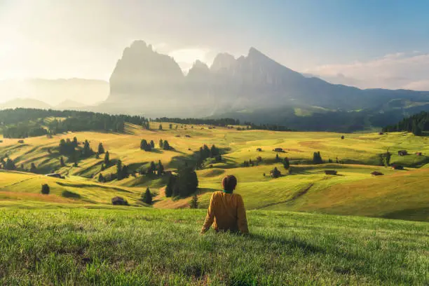 Young man relaxing on alpine meadow in Dolomites Alp mountains Seiser Alm or Alpe di Siusi at sunrise. Trentino Alto Adige or South Tyrol, Italy. Focus on the guy on foreground