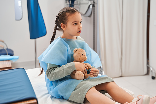 Im afraid. Little cute girl sitting at the hospital bed and embracing her teddy bear while waiting for the procedure. Stock photo
