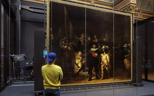 Museum visitor looking at the famous Rembrandt painting, The Night Watch, with some of the equipment used to restore it in the background, at the Rijkmuseum, on Museum Square in Amsterdam
