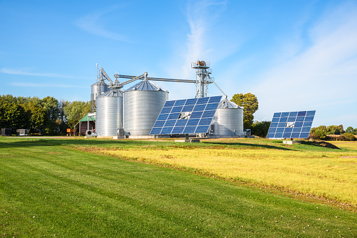 Solar panels in front of a grain elevator with big steel storage bins in a modern farm on a sunny autumn day. Use of renewable energy in agriculture concept. Wolfe Island, ON, Canada.