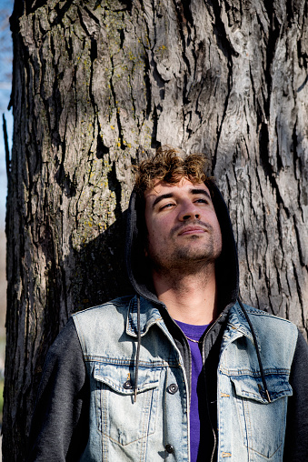 Portrait of young man leaning on a tree. He is in his early thirties, wearing a jean vest and a hoodie. Vertical waist up outdoors shot with copy space.