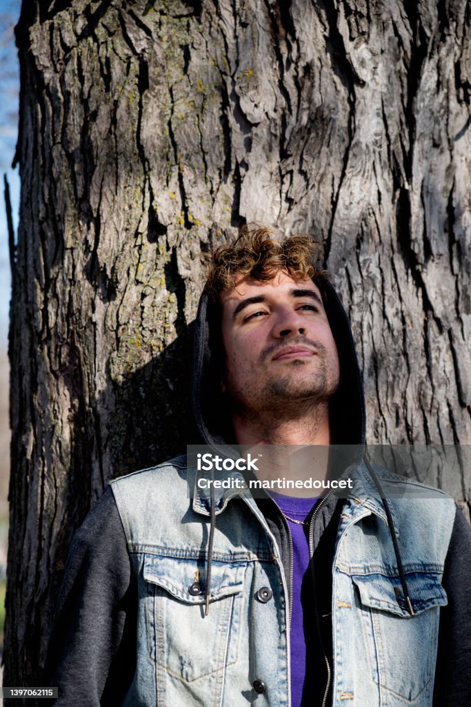 Portrait of young man leaning on a tree. Portrait of young man leaning on a tree. He is in his early thirties, wearing a jean vest and a hoodie. Vertical waist up outdoors shot with copy space. Adult Stock Photo