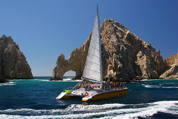 Sailing at Cabo San Lucas Arch A catamaran sails past the landmark Los Arcos rock formation at Land's End in Cabo San Lucas, Mexico. cabo san lucas stock pictures, royalty-free photos & images