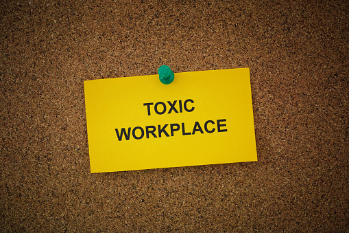 A sticky note with the words Toxic Workplace on it pinned to a cork board. Close up.