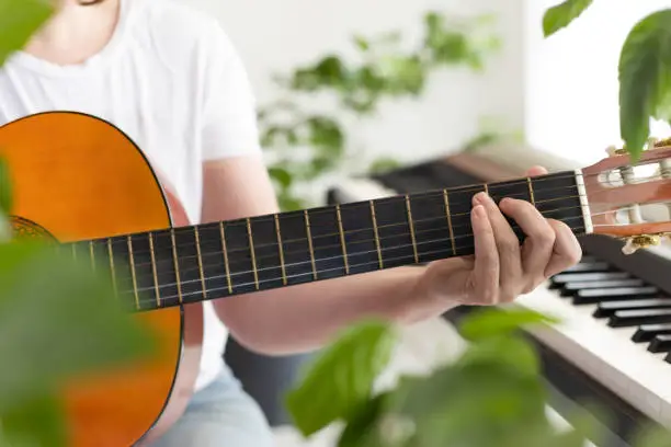 Women's hands play the guitar. Fingers pinch the strings on the fretboard. Home music making, relaxation. Biophilic interior design. Lots of houseplants. Natural daylight.