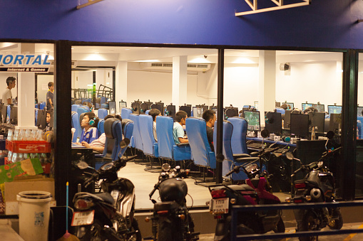 Young thai men are sitting inside of gaming shop at pc. View into shop at night. Shop is near CRU university in Bangkok Chatuchak. Many young men are inside