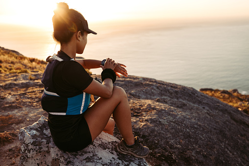 Woman hiker sitting on a rock on the top of the mountain checking smartwatch. Female in sportswear on a hill at sunset.