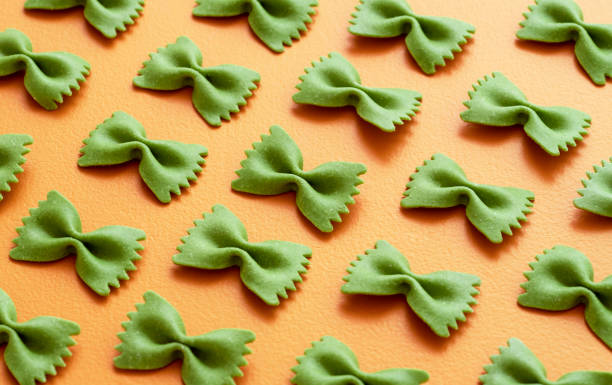 Green farfalle pasta on an orange table. Uncooked spinach pasta Spinach farfalle pasta aligned symmetrically on an orange table. Close-up with uncooked bow tie-shaped pasta. spinach pasta stock pictures, royalty-free photos & images
