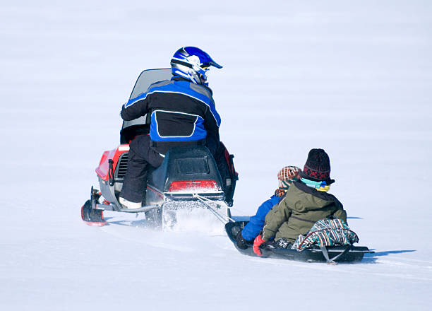 A man on a snowmobile pulling two small children behind him stock photo