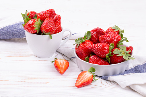 Juicy strawberries berries in a white ceramic bowl and cup on a white shabby wooden table in a rustic style, copy space.