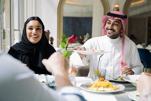 Over the shoulder view of man in dish dash, kaffiyeh, and agal sitting beside woman in abaya and hijab, both laughing as they offer food to friend.