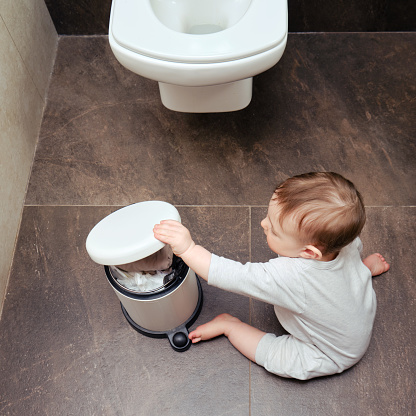 Toddler baby boy is playing in the toilet room with a trash can. Child plays on a brown floor in a beige bathroom with a trash can lid