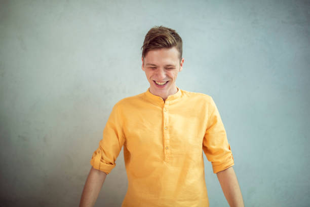 A man in a blouse stands in front of a white background and laughs.