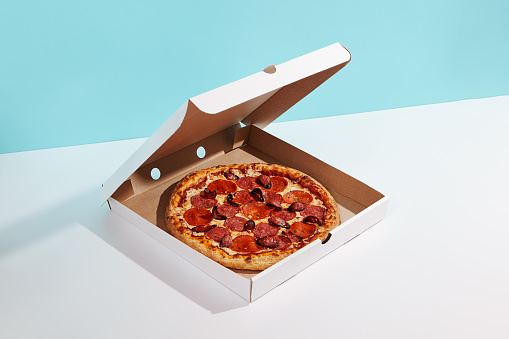 Italian pizza in paper box on coloured background. Meat pizza with sausage and salami in delivery box in minimal style . American pizza delivery concept with color backdrop