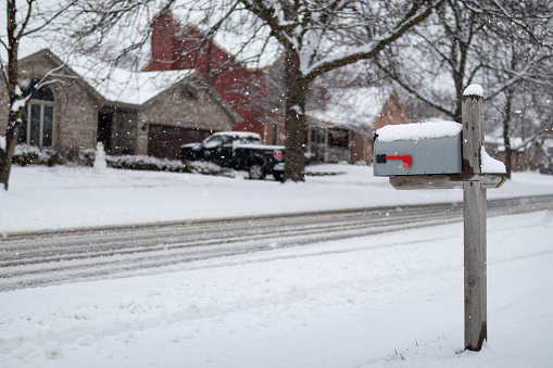 A simple home mailbox along a snow covered neighborhood street with homes during a beautiful snowfall in suburban Illinois