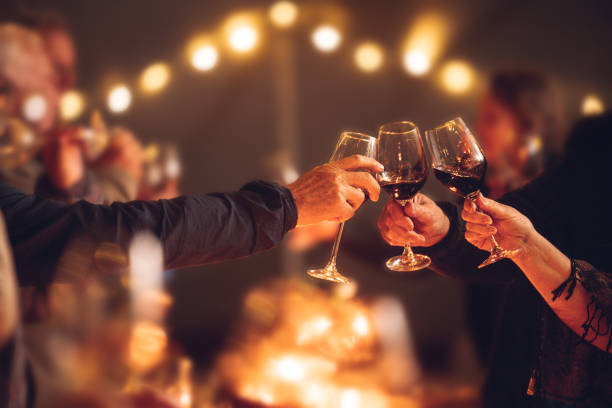 Celebratory red wine toast between senior adult friends at candle light social event party with string fairy lights Celebratory red wine toast between senior adult friends at candle light social event party with string fairy lights office parties stock pictures, royalty-free photos & images