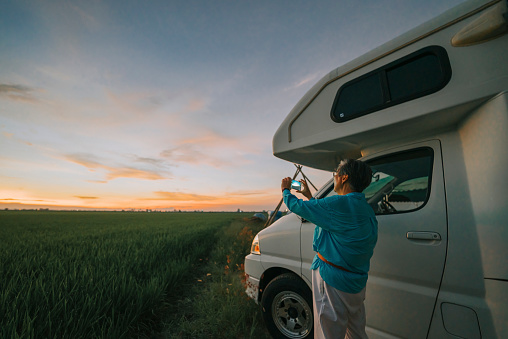 Asian chinese senior woman photographing sunset at paddy field with smart phone beside campervan motor home