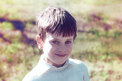 Grainy analog portrait of an elementary-aged boy looking at camera outdoors. Vintage photo from the seventies.