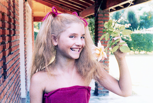 Analog photo of a blonde young girl smiling and looking at the camera while showing a rose flower. Grainy image from the eighties.