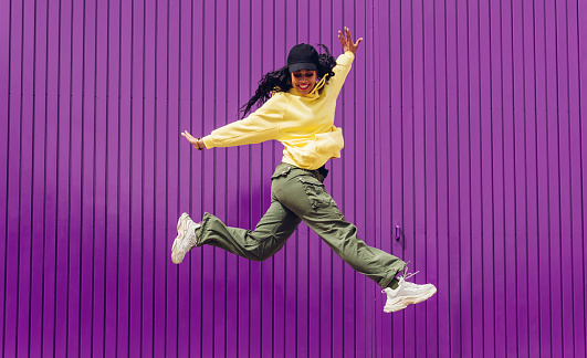 Jumping young woman in yellow dress on purple background. Dancer of hip hop and trap music. Concept of freedom, celebration, joy.